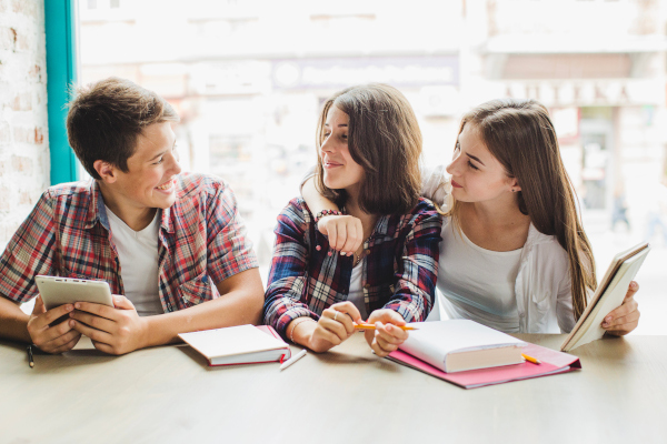 Adolescentes charlando sonrientes en clase con una tablet, cuadernos y libros